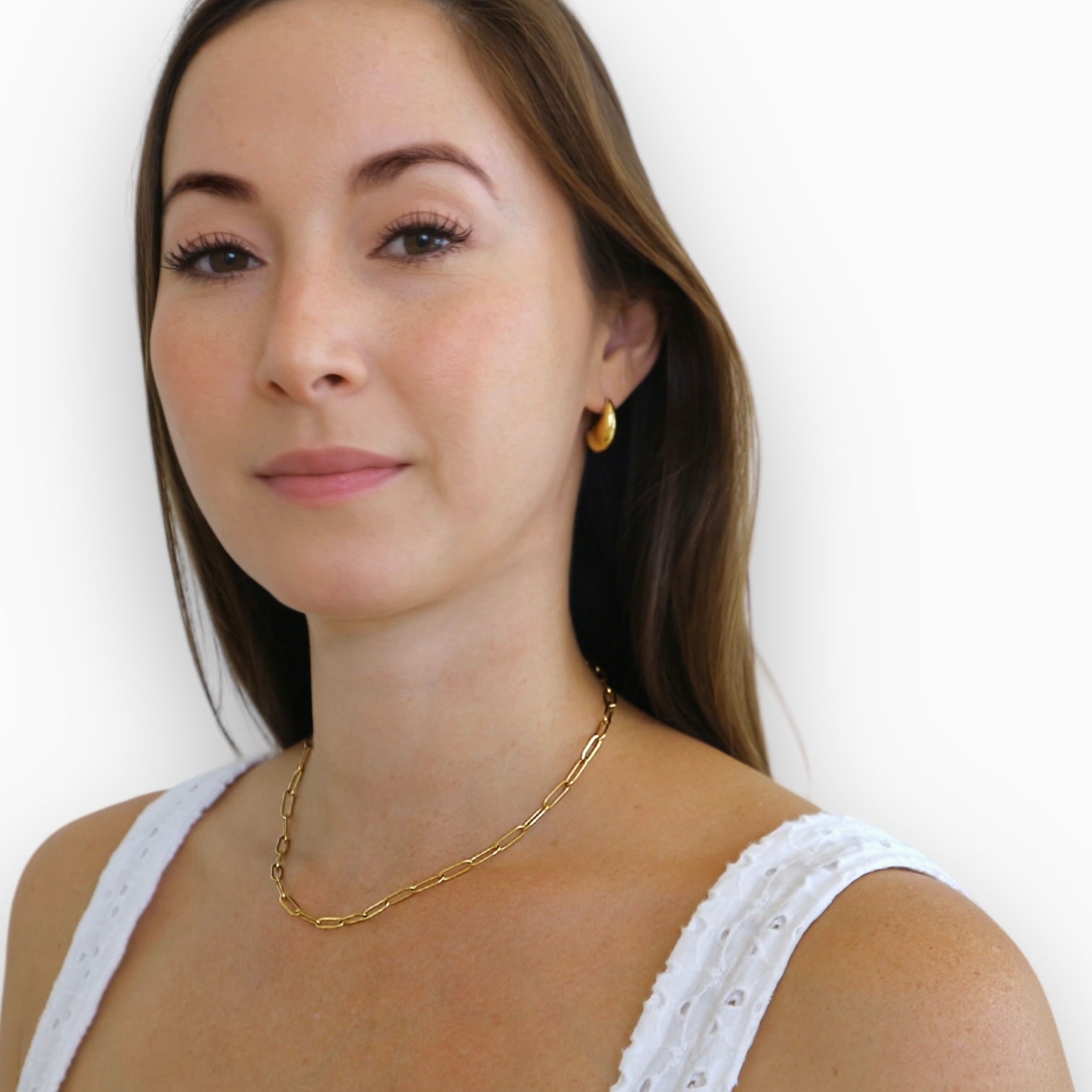 brunette girl white background. wearing gold earrings and paperclip necklace. white tank