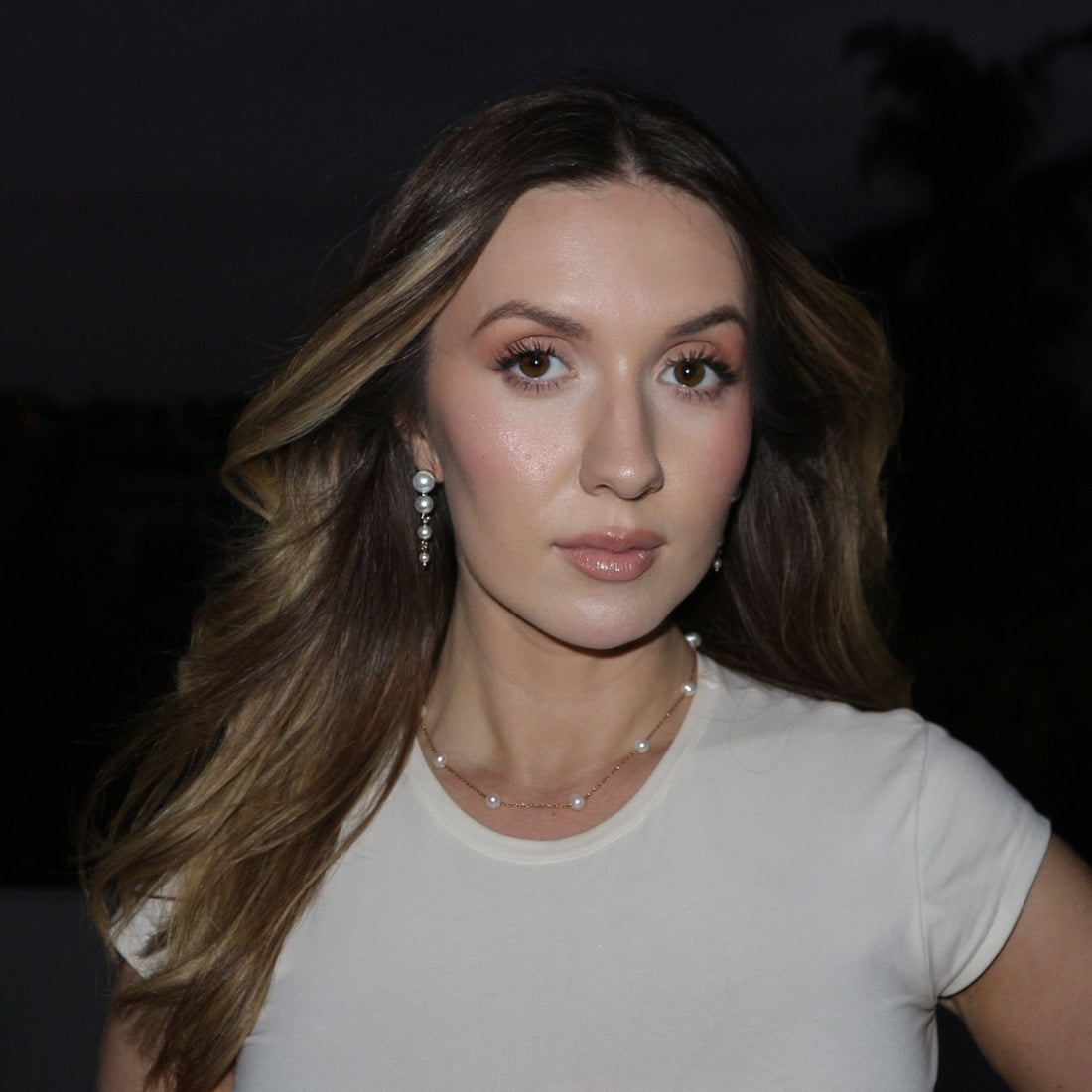 Woman with long brown hair wearing a white top against a dark background