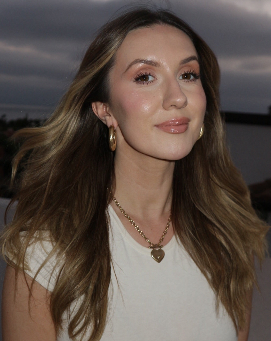 Woman with long brown hair and a white top standing outdoors with a cloudy sky.